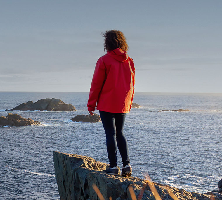 Woman in red jacket is standing at the edge of a cliff enjoying the beautiful ocean scenery.