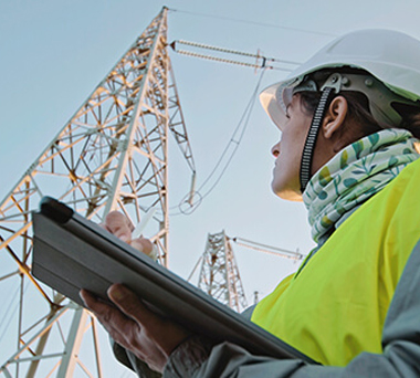 Woman in safety gear with tablet looking up electrical tower