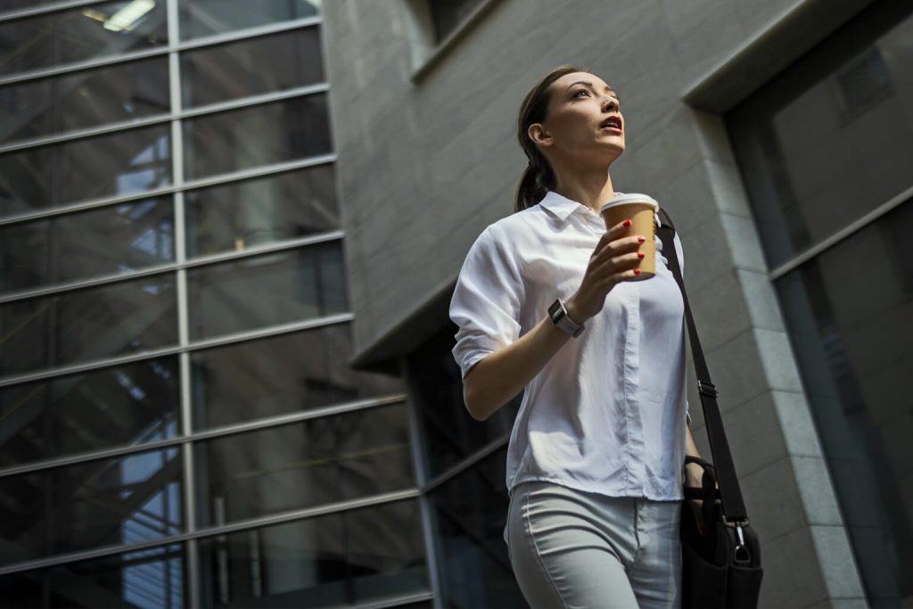 Casually-dressed business woman walking a city street holding a coffee cup and looking up
